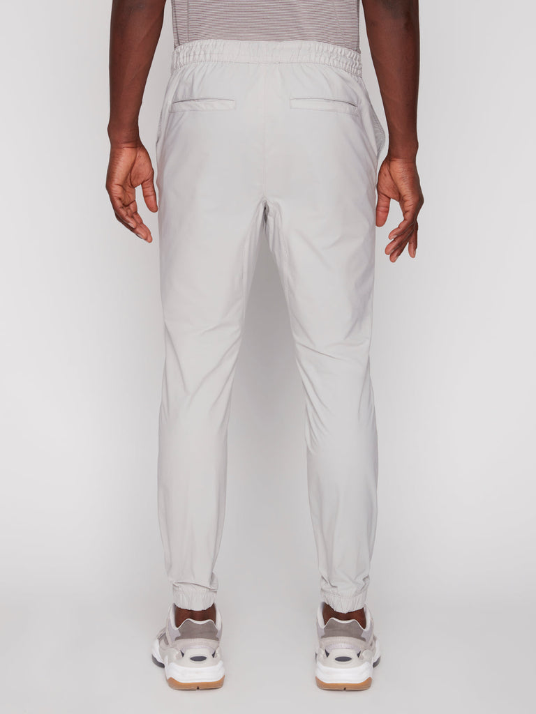 A man faces away from the camera, wearing Projek Raw’s TECH JOGGER - BONE with elastic cuffs, paired with white sneakers and a gray shirt. His arms hang relaxed by his sides against a plain white background.