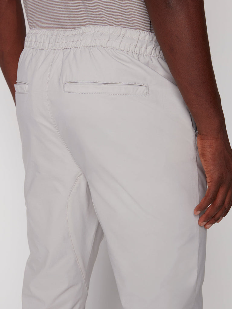 Close-up of the back of a person wearing Projek Raw TECH JOGGER - BONE, featuring light gray color, elastic waistband, and two back welt pockets; their right arm rests at their side.