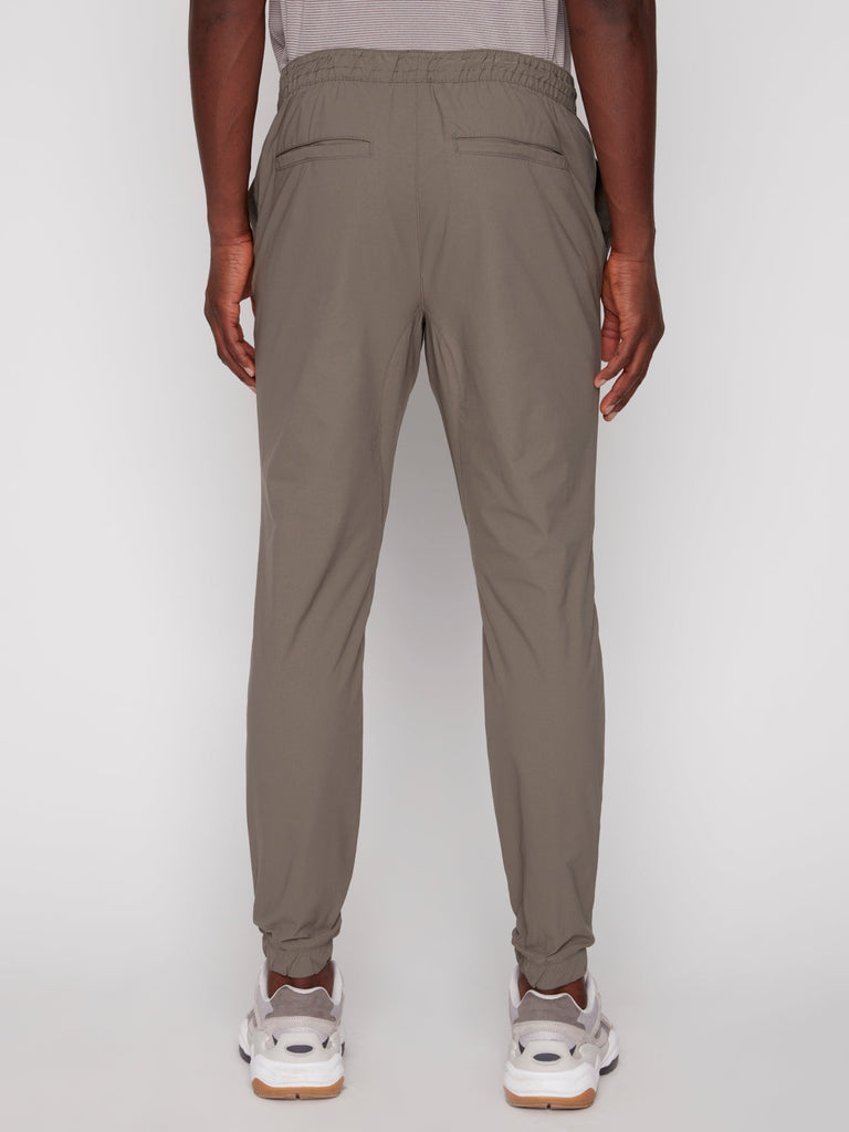 A person wearing Projek Raw TECH JOGGER - MOSS stands with their back to the camera against a white background. The light gray joggers feature an elastic waistband and back pockets, paired with white sneakers.
