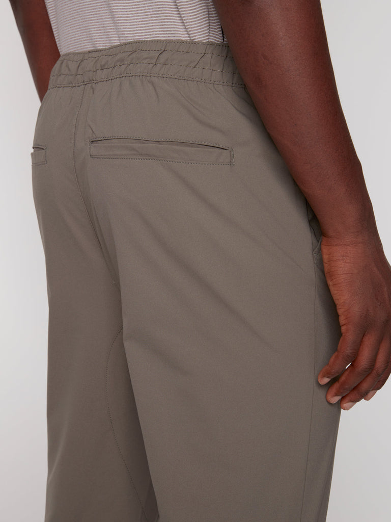 A close-up of a person wearing Projek Raw TECH JOGGER - MOSS, featuring an elastic waistband and welt back pockets. Their right hand is near the side seam, paired with a gray and white striped shirt.