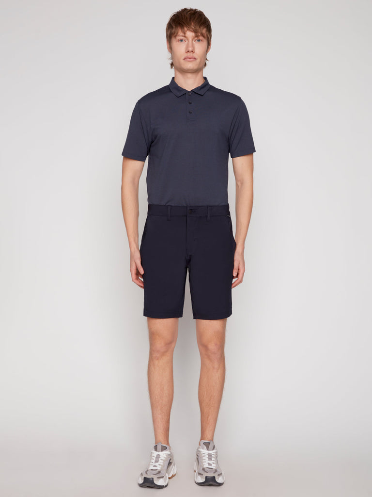 A man stands against a plain light background, wearing Projek Raw’s DAMIAN MENS SHORT in navy, a dark navy short-sleeve polo shirt, and light gray athletic shoes.