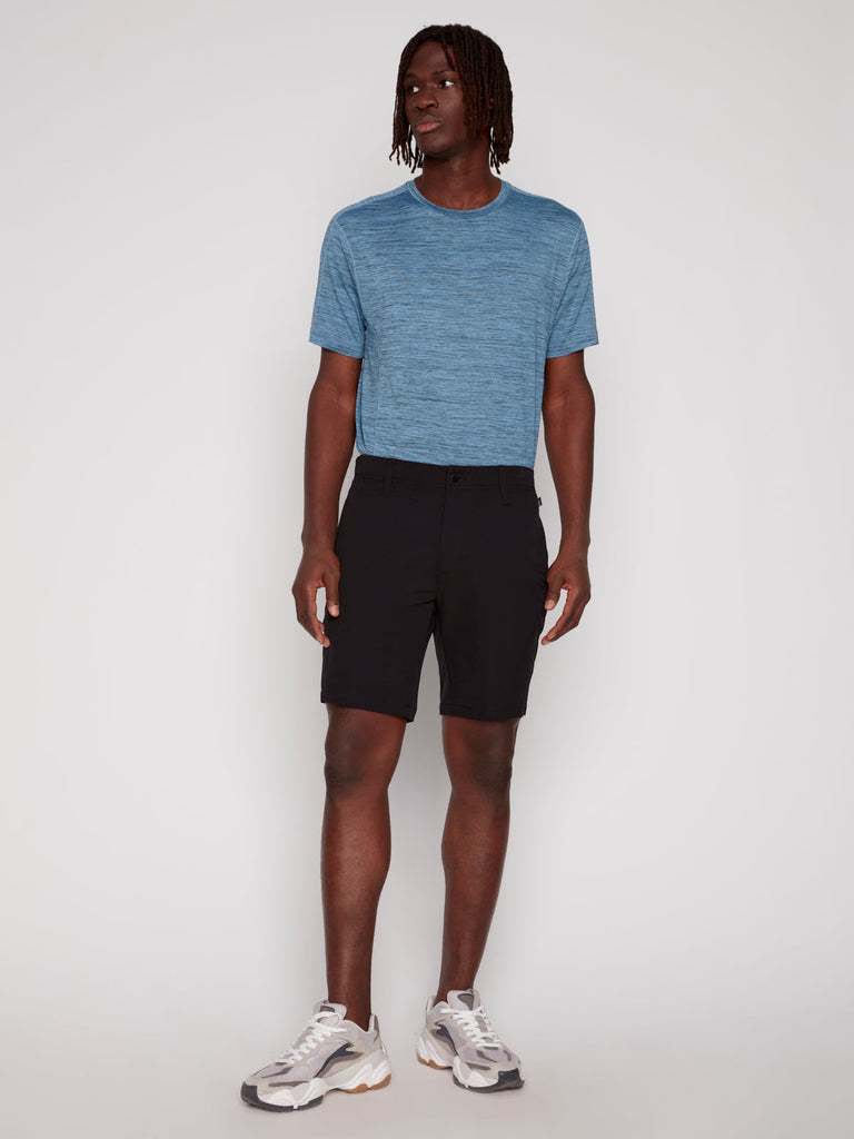 A person stands against a plain light background wearing the Projek Raw ANTHONY MENS SHORT - BLACK, paired with a blue short-sleeve shirt and white sneakers with gray and tan accents, looking slightly to the side in a relaxed posture.