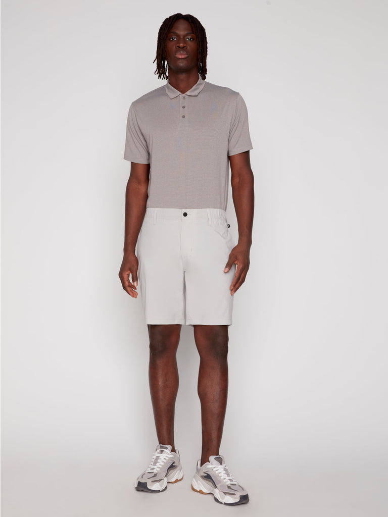 A man stands facing forward against a plain background, wearing Projek Raw ANTHONY MENS SHORT - BONE, a light gray short-sleeve polo shirt, and white sneakers with gray and beige accents.