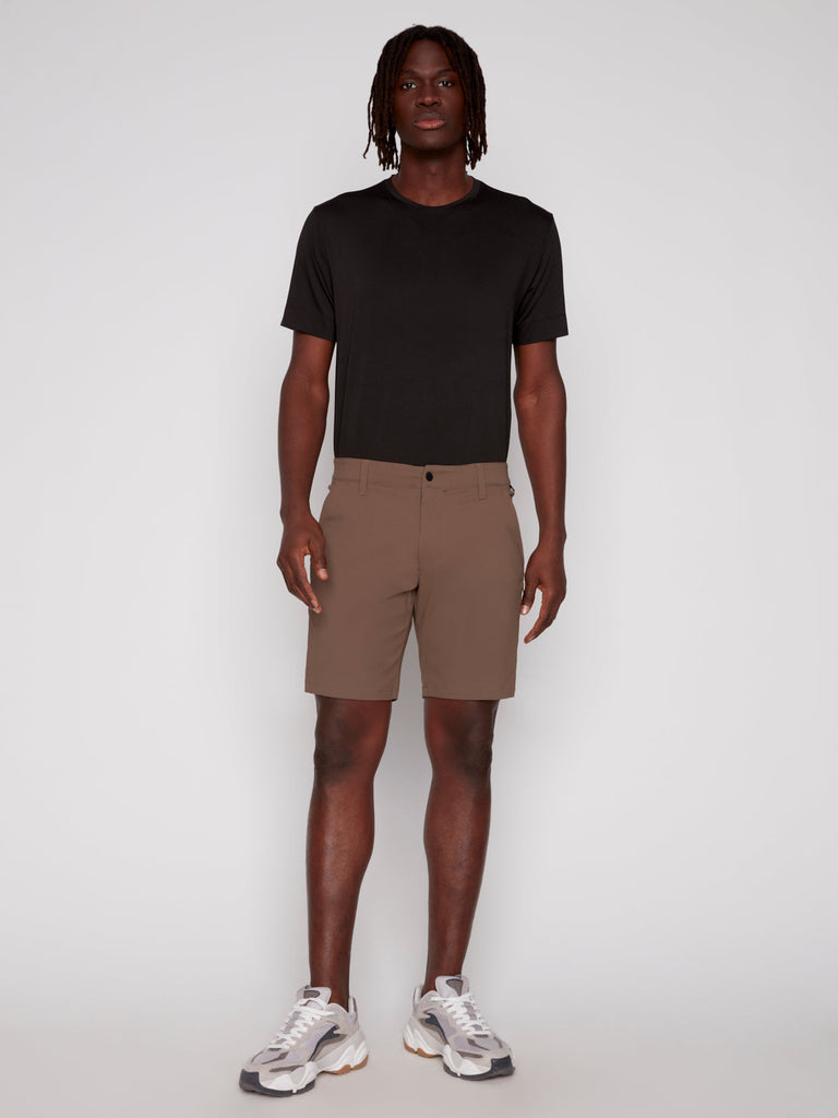 A person stands against a light gray background, facing forward in a neutral pose, wearing Projek Raw’s PAUL MENS SHORT - COFFEE, a black short-sleeve t-shirt, and white sneakers with gray accents.