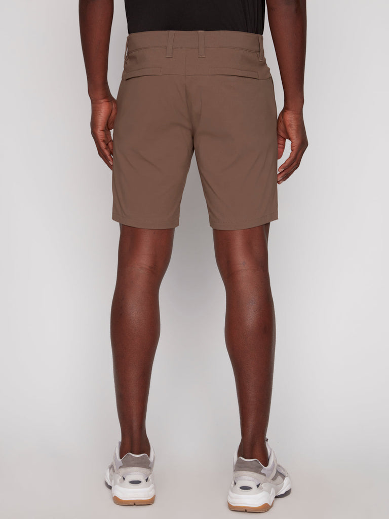 A man wearing Projek Raw’s PAUL MENS SHORT in Coffee, a black shirt, and white athletic shoes stands facing away from the camera against a plain light background.