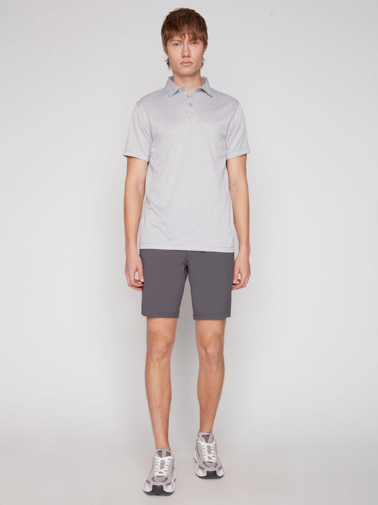 A young man stands facing forward against a plain light background, wearing the Projek Raw MICHAEL MENS POLO in light grey, paired with dark gray shorts and gray sneakers.