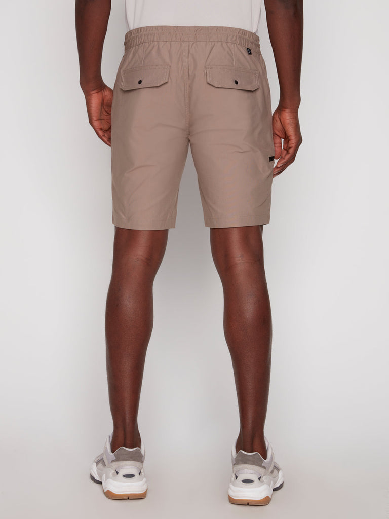 A person is shown from behind wearing Projek Raw LUKA MENS SHORTS - BEIGE and a white t-shirt with white sneakers, standing against a plain white background.