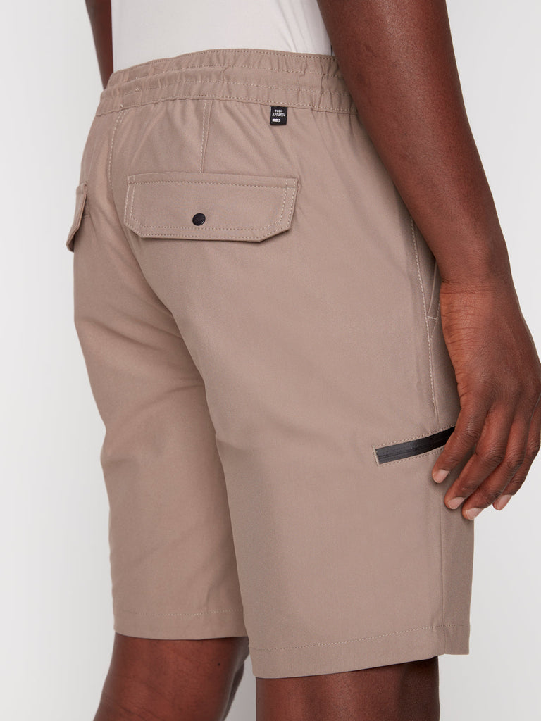 A person is wearing Projek Raws LUKA MENS SHORTS in beige, featuring visible stitching, a buttoned flap back pocket, a small black tag, and back pockets; shown standing sideways with their right hand by their leg.