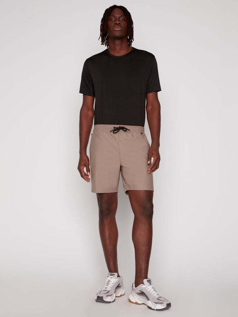 A man stands against a plain background wearing Projek Raws JULIUS MENS SHORT in beige, paired with a black t-shirt and white sneakers with gray and beige accents. He faces forward with a neutral expression and relaxed posture.