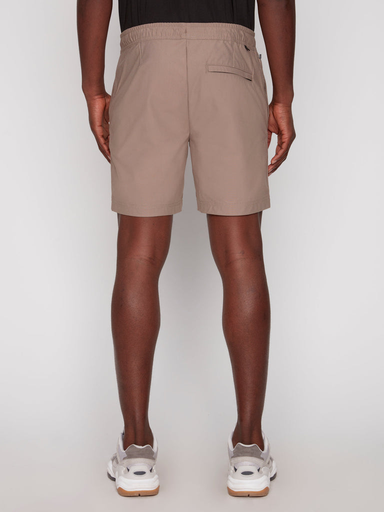 A person faces away, wearing Projek Raw JULIUS MENS SHORT - BEIGE featuring an elastic waistband and back pocket, paired with a black shirt and white sneakers against a plain background.