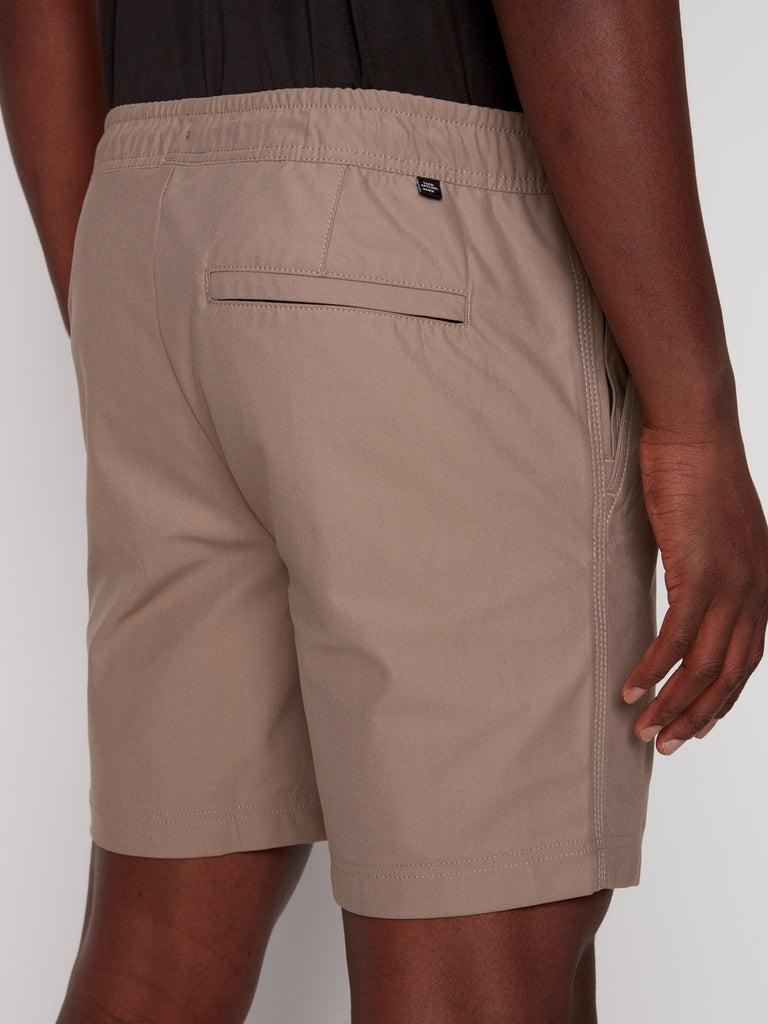 A person wearing Projek Raw JULIUS MENS SHORT in beige, featuring an elastic waistband, side pockets, a back pocket, and a relaxed fit. Their arm is visible alongside their body.