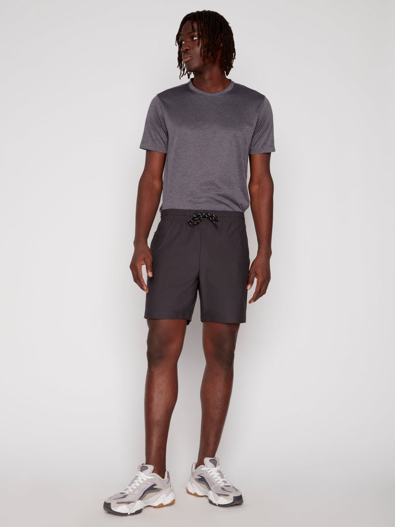 Against a plain background, a person wears the Projek Raw JULIUS MENS SHORT - CHARCOAL paired with a dark grey tee and chunky white sneakers with beige accents, looking to the side with one hand relaxed by their side.
