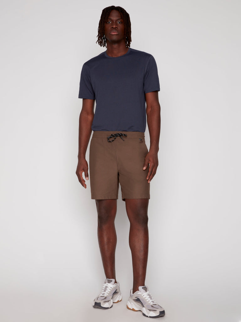 A man stands against a plain background, wearing the Projek Raw JULIUS MENS SHORT in taupe, a navy blue T-shirt, and white and gray sneakers. He faces forward with a neutral expression and relaxed posture.