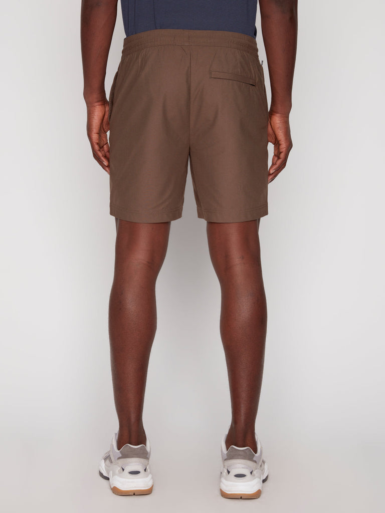 A person faces away, wearing Projek Raws JULIUS MENS SHORT in taupe with an elastic waistband and right back pocket, paired with a dark blue shirt and white sneakers. The background is plain white.