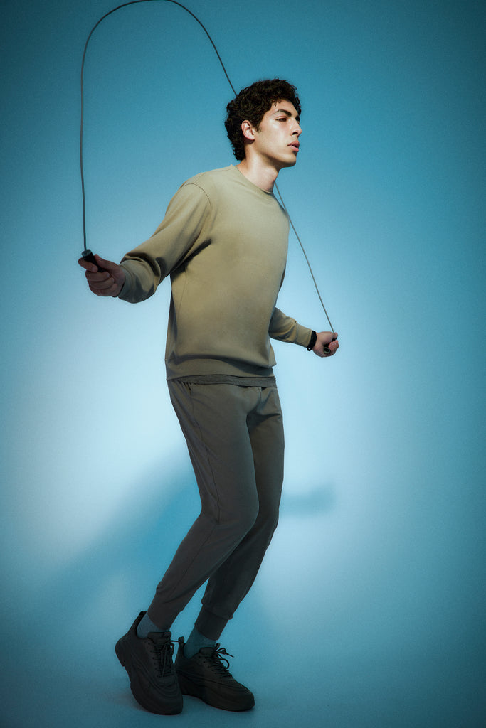 A young man in beige athletic wear is jumping rope against a blue gradient background, holding the handles and mid-jump with a focused expression.