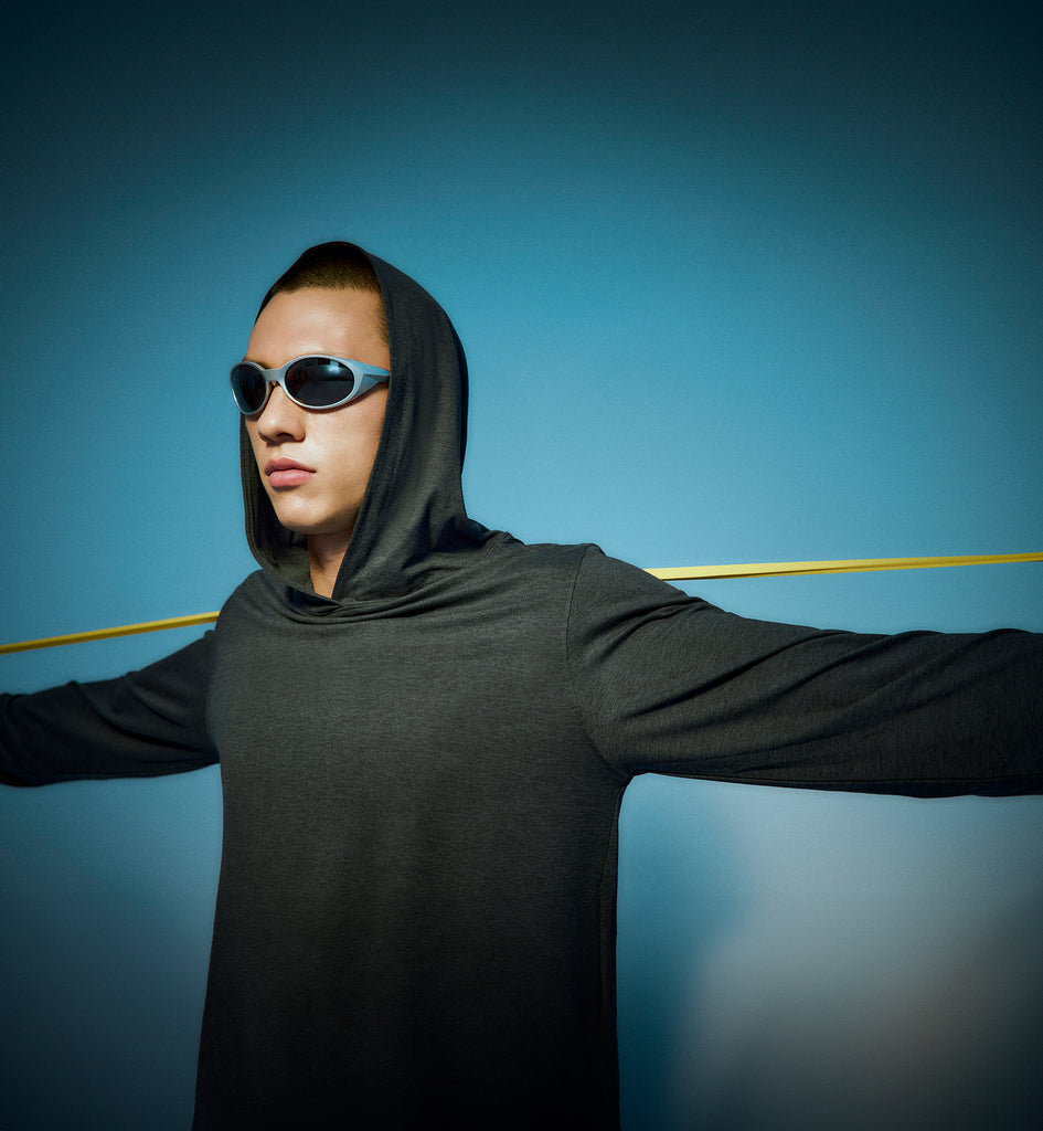 A person wearing a gray hooded shirt and dark sunglasses stands with arms outstretched against a blue background, with a yellow rod positioned behind their shoulders.