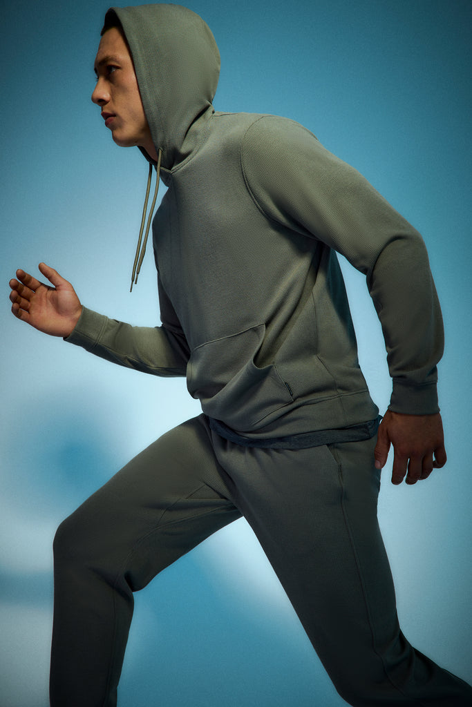 A person in a gray hooded sweatshirt and matching sweatpants poses as if running, against a blue gradient background.