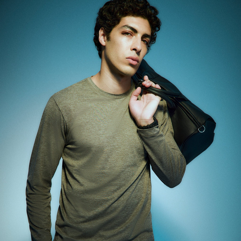 A young man with curly dark hair wears a long-sleeved olive shirt and holds a black bag over his shoulder, standing against a blue gradient background and looking confidently at the camera.