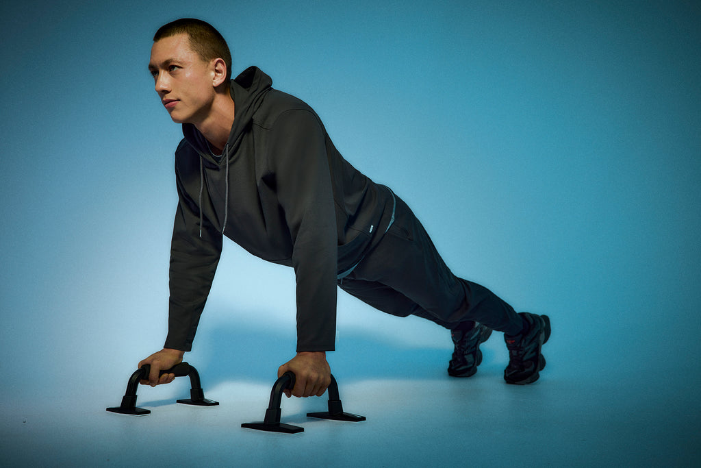 A man in a dark tracksuit does push-ups using push-up bars on a smooth, blue-lit surface. He looks focused and maintains a straight posture, with his body parallel to the floor.