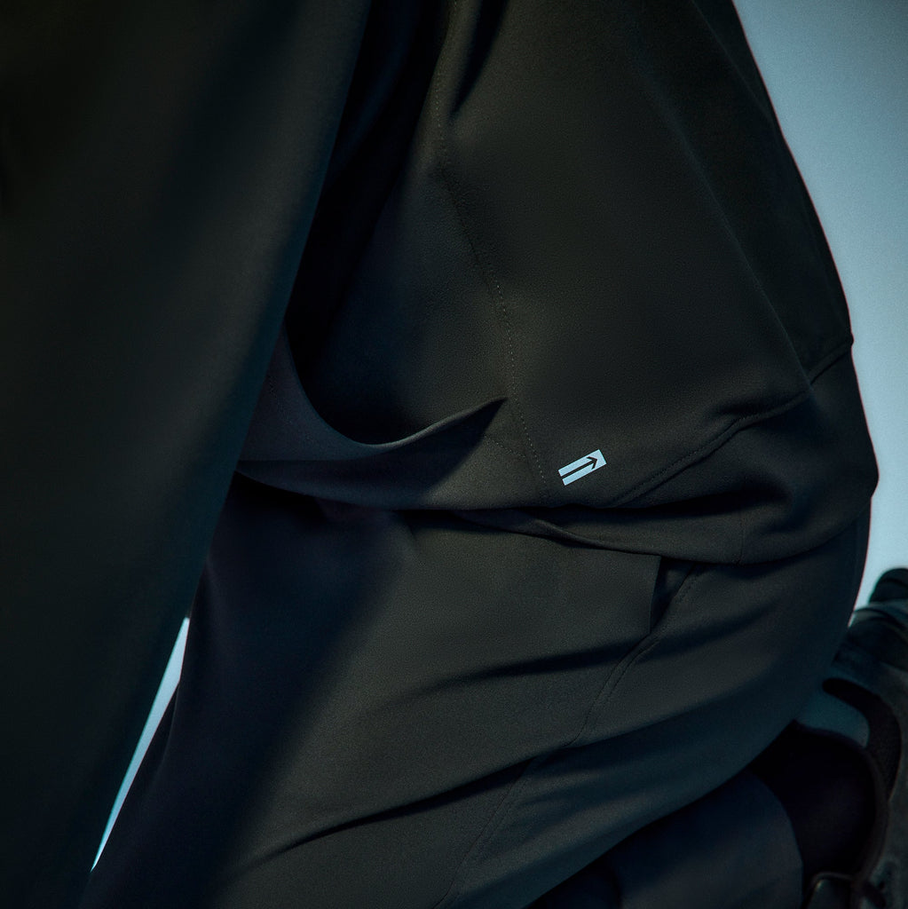 A close-up of a person wearing a black jacket with a small metallic logo near the pocket, showing part of their arm and side against a light background.