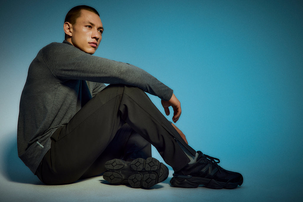 A man in dark athletic wear and black sneakers sits on the floor with his knees drawn up, looking thoughtfully into the distance against a blue gradient background.