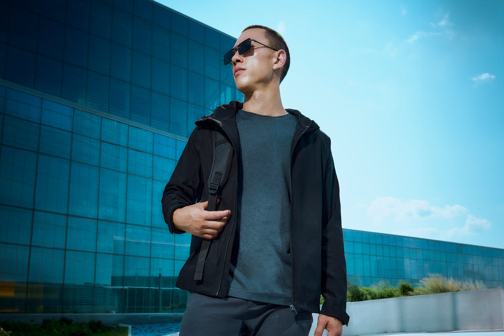 A man wearing sunglasses, a dark jacket, and a t-shirt stands outside a modern glass building, holding a strap of his backpack and looking confidently into the distance under a clear blue sky.