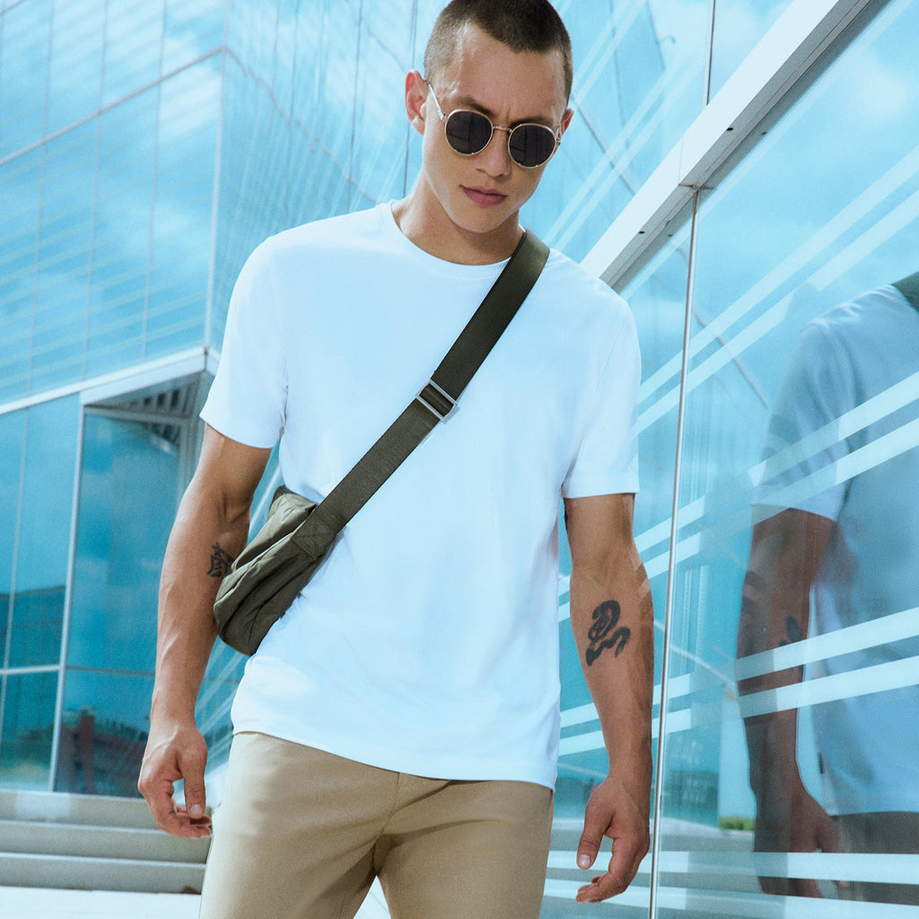 A man wearing sunglasses, a white t-shirt, beige pants, and a crossbody bag walks outdoors near a modern glass building on a sunny day.