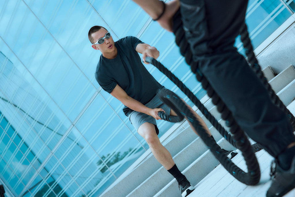 A man wearing sunglasses and sportswear exercises outdoors using battle ropes, with another person holding the ropes in the foreground. They are near modern glass buildings and stairs.