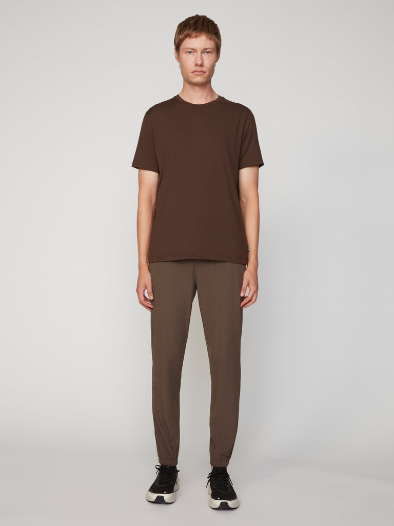 A person stands against a plain background, wearing the TECH APPAREL MEN’S PEACH:TECH CLASSIC FIT CREW NECK T-SHIRT in dark brown, paired with light brown pants and black sneakers.