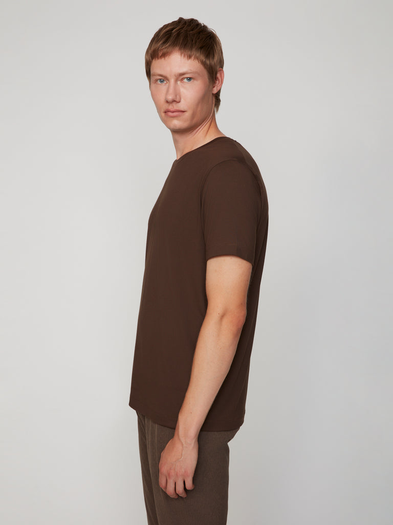 A young man with short light brown hair stands sideways against a light gray background, wearing the TECH APPAREL MEN’S PEACH:TECH CLASSIC FIT CREW NECK T-SHIRT and matching brown pants, looking at the camera with a neutral expression.
