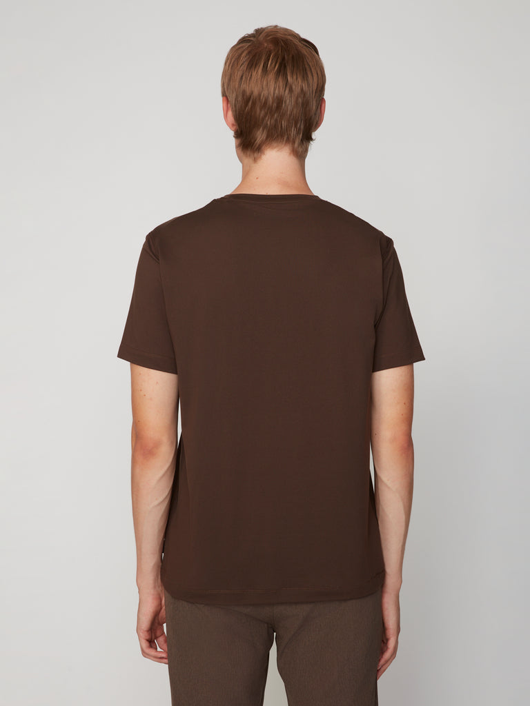 A person with short light brown hair faces away, wearing the TECH APPAREL MEN’S PEACH:TECH CLASSIC FIT CREW NECK T-SHIRT and matching pants in dark brown against a light gray background. The look is simple and performance-driven.