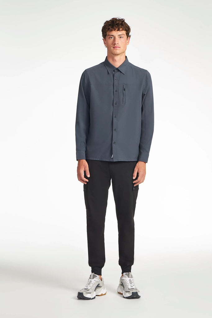 A man stands facing forward against a plain white background, wearing a dark blue button-up shirt, white sneakers, and TECH APPAREL MENS FLUX 2.0 CARGO JOGGER in black stretch flux fabric.