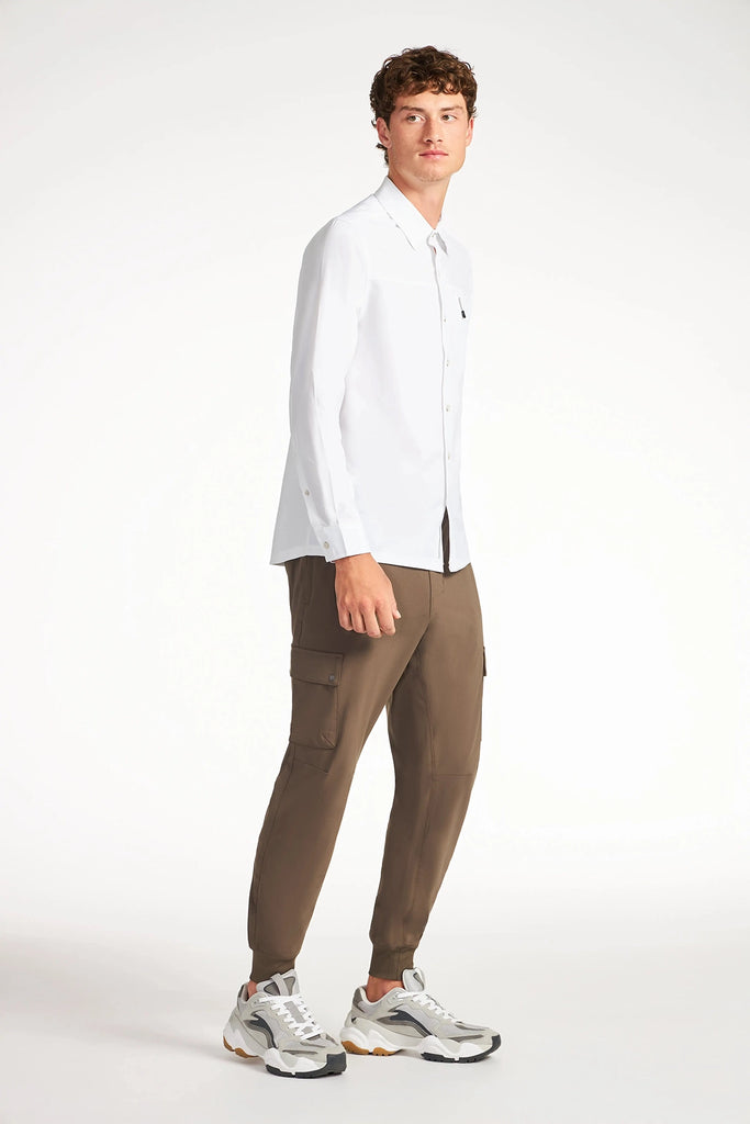 A young man wears TECH APPARELs MENS FLUX 2.0 CARGO JOGGER in brown stretch flux fabric with a white long-sleeve shirt and light sneakers, standing against a plain white background, looking slightly left with a neutral expression.