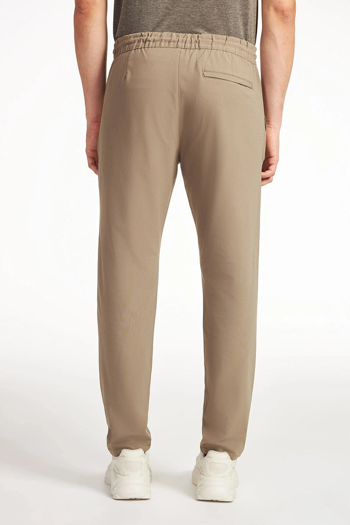 A person is seen from the back wearing TECH APPARELs MENS FLUX 2.0 PANT WITH DRAWCORD WAIST in light brown, paired with a gray t-shirt and white sneakers, set against a plain white background.