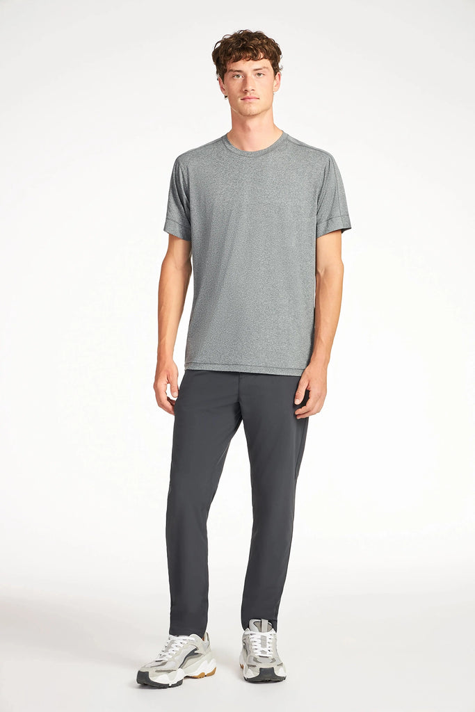 A young man with short brown hair wears a gray tee, white sneakers, and TECH APPARELs MENS FLUX 2.0 PANT WITH DRAWCORD WAIST, offering a classic fit and modern comfort, standing against a plain white background with a neutral expression.
