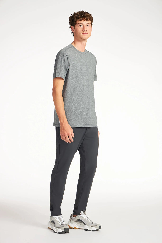 A young man stands against a white background, wearing TECH APPAREL’s MENS FLUX 2.0 PANT WITH DRAWCORD WAIST, a gray short-sleeve tee, and white sneakers. He faces slightly left with a relaxed expression.