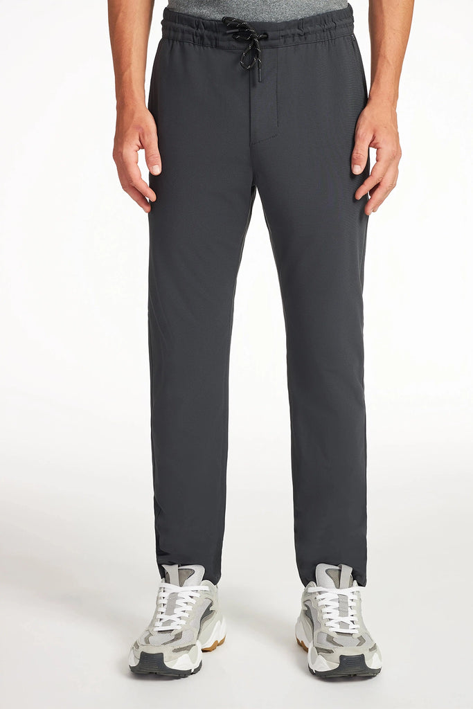 A person wearing TECH APPAREL MENS FLUX 2.0 PANT WITH DRAWCORD WAIST in dark gray and light gray sneakers stands against a white background, with only the lower torso and legs visible.