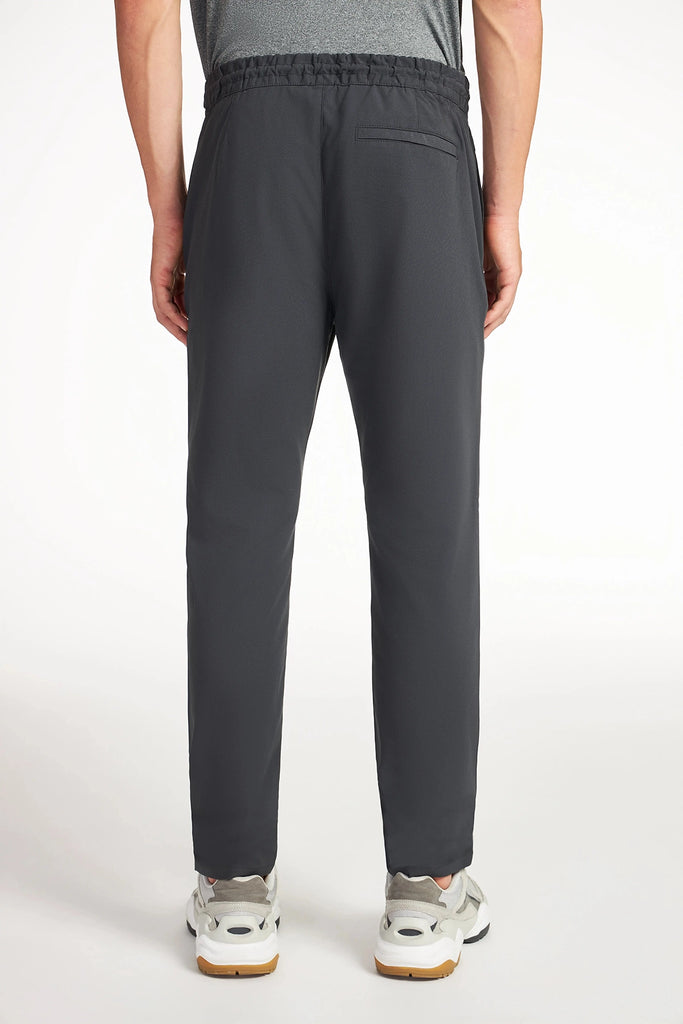 A person is shown from the back wearing TECH APPAREL’s MENS FLUX 2.0 PANT WITH DRAWCORD WAIST in dark gray with white sneakers, standing against a plain white background.