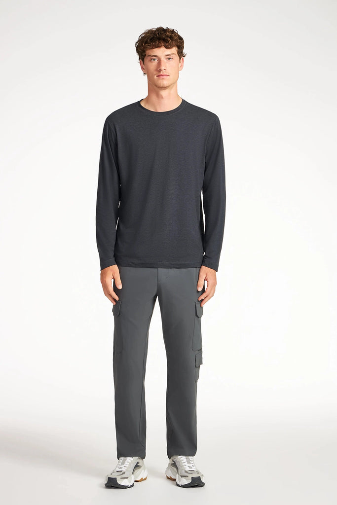 A young man faces forward against a white background, wearing TECH APPAREL’s MENS DRI-FLEX CARGO PANT, a long-sleeve dark gray shirt, and white sneakers.