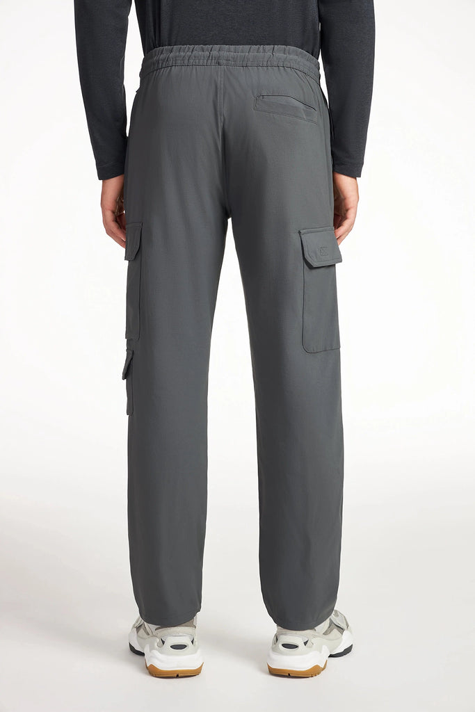 A person faces away, wearing TECH APPAREL MENS DRI-FLEX CARGO PANT in gray with multiple pockets, elastic waistband, and a casual fit, paired with white sneakers. The wrinkle-resistant pants are shown against a plain white background.