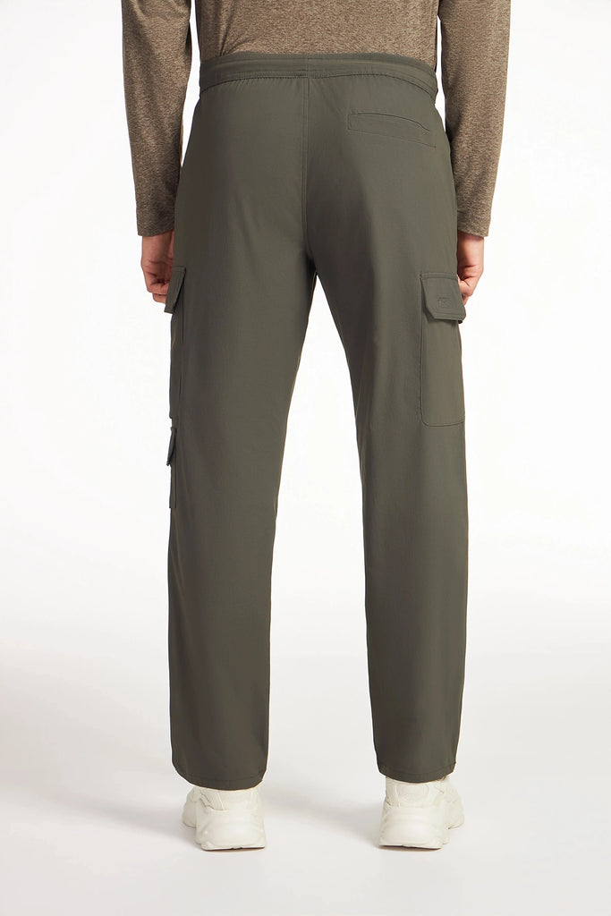A person stands facing away, showcasing the TECH APPAREL MENS DRI-FLEX CARGO PANT in olive green with utility cargo pockets, paired with a brown long-sleeve shirt and white shoes against a plain background.