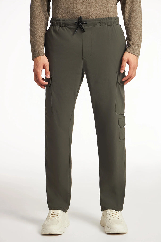 A person wears TECH APPAREL MENS DRI-FLEX CARGO PANT in olive green, styled with utility cargo pockets and a drawstring waist, paired with a brown long-sleeve shirt and white sneakers, standing against a plain white background.