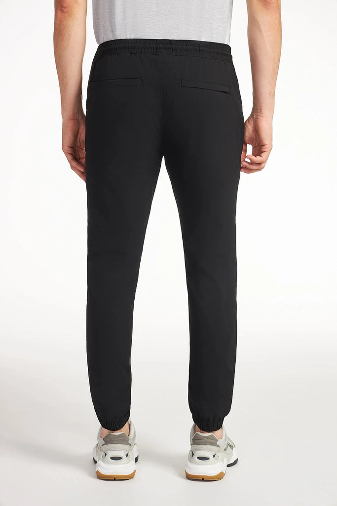 A person in TECH APPAREL MENS DRI-FLEX JOGGER and white sneakers stands with their back to the camera against a plain white background. The jogger features elastic cuffs, an athletic fit, and two back pockets.