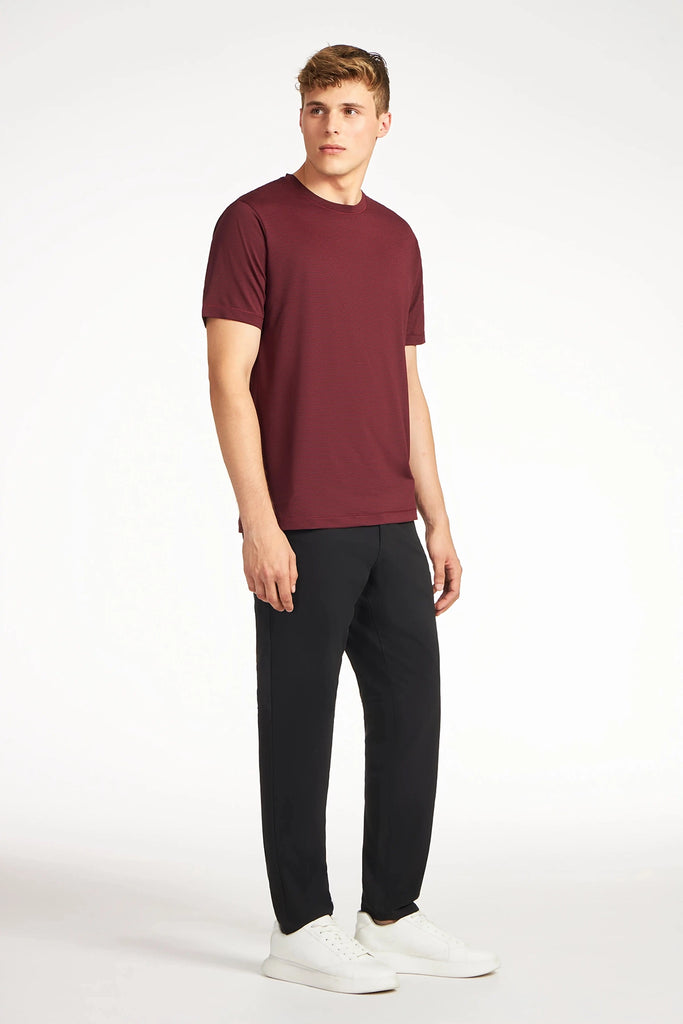 A young man stands against a plain white background, wearing a maroon short-sleeve t-shirt, TECH APPAREL MENS DRI-FLEX PANTS, and white sneakers. He looks slightly to his left with a neutral expression.