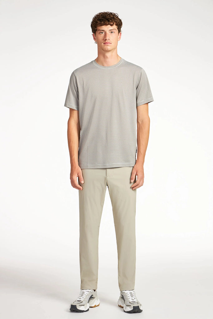 A young man with short curly brown hair stands facing forward in a light gray t-shirt, TECH APPAREL MENS DRI-FLEX wrinkle-resistant beige pants, and white sneakers against a plain white background.