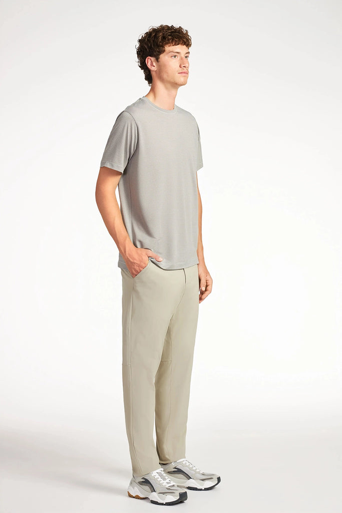 A young man with curly hair faces right, wearing a light gray t-shirt, TECH APPAREL MENS DRI-FLEX PANTS, and gray sneakers against a plain white background.