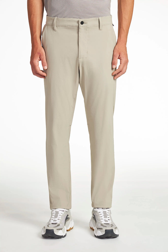 A model wears TECH APPAREL MENS DRI-FLEX PANTS in light beige, styled with gray and white sneakers and a tucked-in gray shirt, against a white background. The image emphasizes the modern fit of the wrinkle-resistant pants.