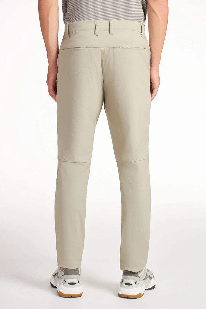 A person models TECH APPAREL MENS DRI-FLEX PANTS in light beige, standing with their back to a plain white background. These slim-fit, wrinkle-resistant pants have a seam above the knee and are styled with white sneakers for a modern look.
