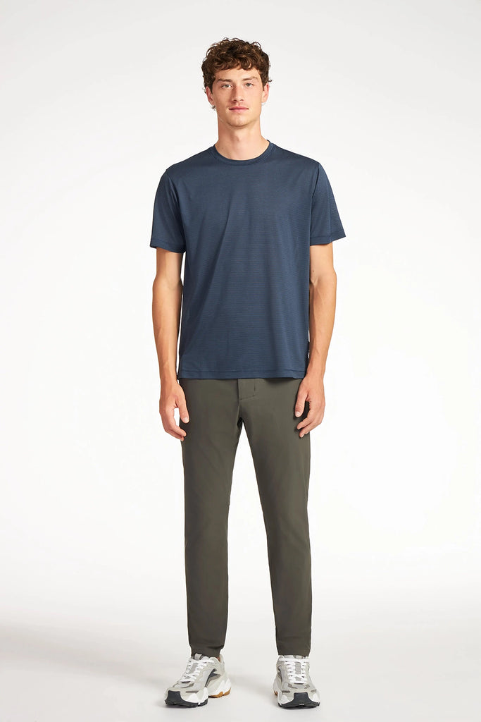 A young man stands facing forward, wearing a dark blue t-shirt, TECH APPAREL MENS DRI-FLEX PANTS in olive green, and white sneakers against a plain light background.