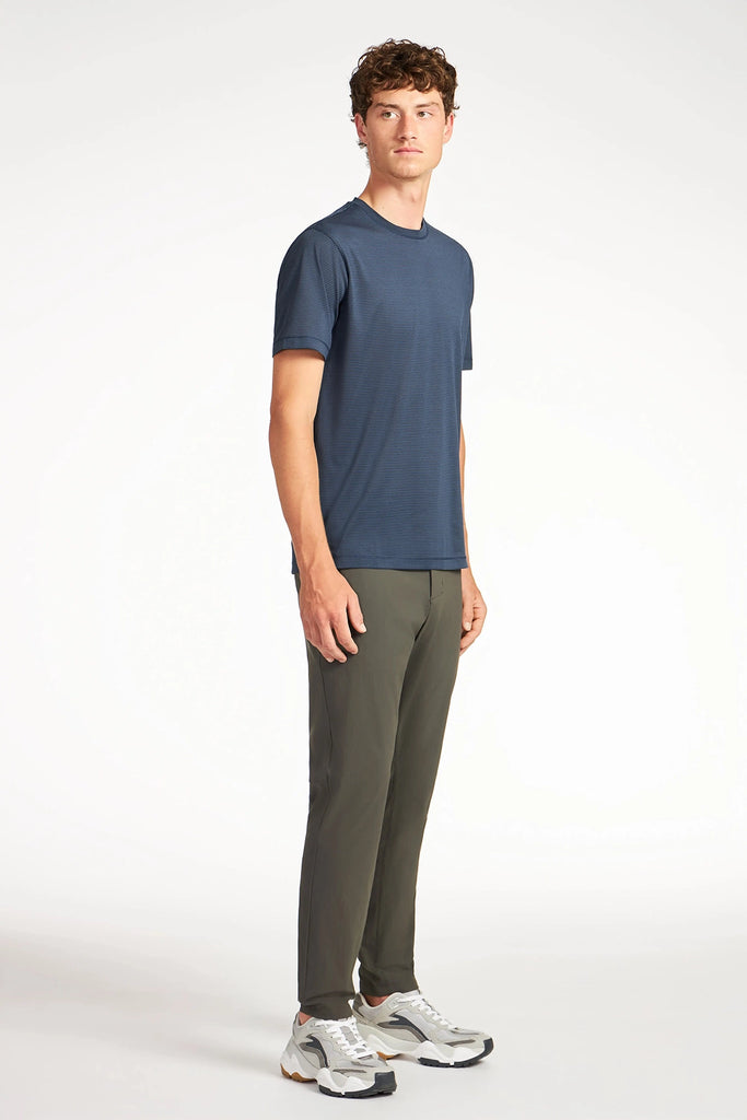 A man faces slightly to the side, wearing a navy blue t-shirt, TECH APPAREL MENS DRI-FLEX PANTS in olive green made from breathable fabric, and gray sneakers, set against a plain white background.